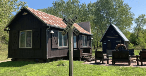 A rustic cabin with a metal roof, surrounded by greenery, next to a railroad crossing sign and outdoor seating area.
