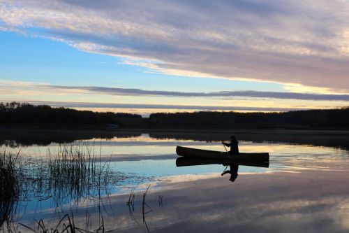 A person paddles a canoe on a calm lake at sunset, with reflections of clouds and trees in the water.