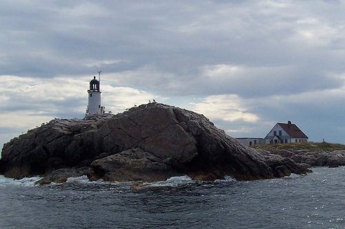 A lighthouse on a rocky island with a house nearby, under a cloudy sky and calm waters.