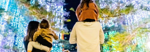 A family enjoys a festive light display, with one child on an adult's shoulders, surrounded by colorful lights.