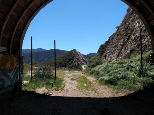 View from an archway into a mountainous landscape, with a dirt path leading through greenery under a clear blue sky.