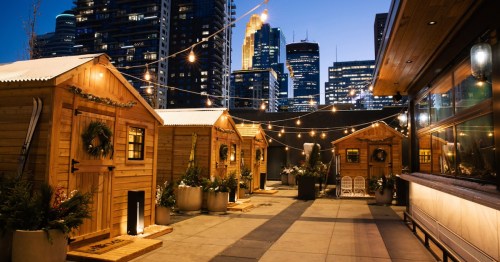 Cozy wooden cabins with festive decorations, string lights, and a city skyline at dusk in the background.