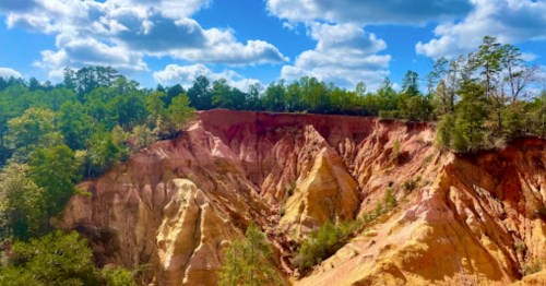 A colorful canyon with layered rock formations under a blue sky with fluffy clouds and green trees in the background.