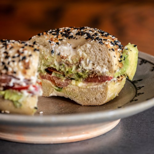 A close-up of a sesame bagel with avocado, tomato, and cream cheese, partially bitten, on a decorative plate.