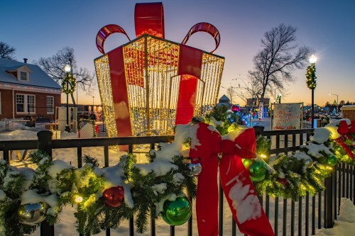 A large, illuminated gift box surrounded by festive decorations and snow, with a sunset in the background.