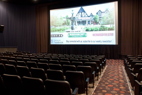 Interior of a movie theater with empty black seats facing a large screen displaying promotional content.