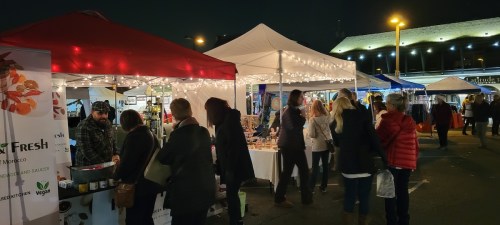 A bustling night market scene with vendors under tents, string lights, and shoppers browsing various stalls.