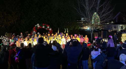 A large group of performers in festive attire sings on stage, surrounded by a crowd and holiday decorations at night.