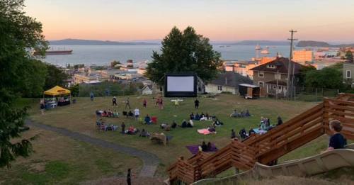 Outdoor movie screening on a grassy hill, with people seated on blankets, overlooking a waterfront at sunset.