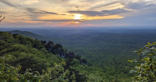 A scenic view of rolling green hills under a colorful sunset, with a vast sky and distant mountains in the background.