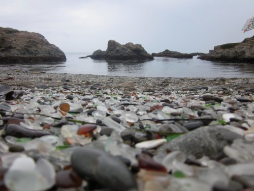 A beach covered in colorful sea glass, with rocky formations and calm water in the background under a cloudy sky.