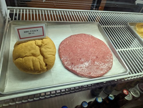 A large, round yellow bun labeled "Big Ugly" next to a flat, pink meat patty on a display tray.