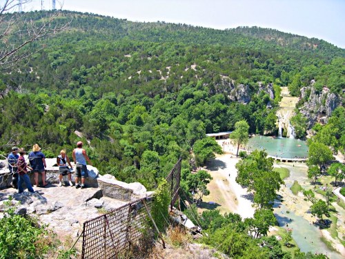 A scenic view from a rocky overlook, showing a lush green landscape with a pond and waterfall below.