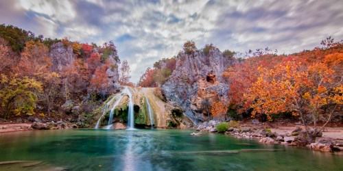 A serene waterfall cascades into a clear pool, surrounded by autumn foliage and rocky cliffs under a cloudy sky.