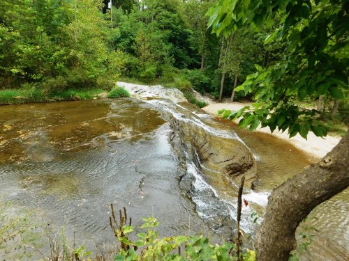 A serene river flows over a rocky ledge, surrounded by lush greenery and trees.