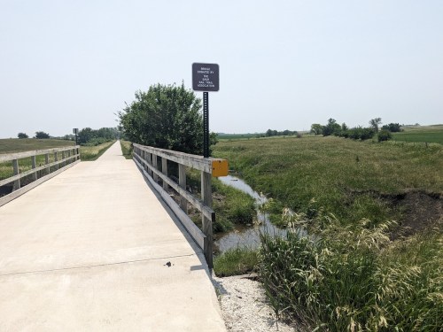 A wooden bridge over a small stream, with grassy fields and a sign in the background on a sunny day.