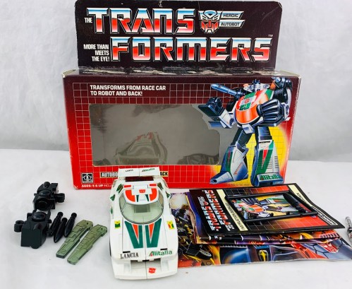 A vintage Transformers toy, a white race car with green and red details, alongside its box and accessories.
