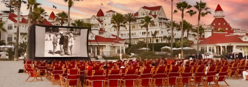 Outdoor movie screening on a beach, with rows of chairs facing a large screen and a historic hotel in the background.