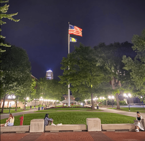 A night scene of a park with an American flag, trees, and people sitting on benches.