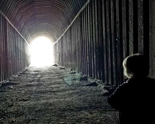 A person stands in a dark tunnel, facing a bright light at the end, creating a sense of depth and mystery.