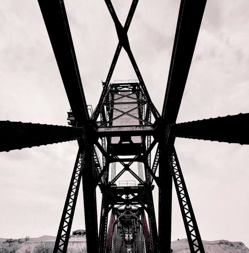 Black and white image of a bridge's intricate metal structure viewed from below, against a cloudy sky.