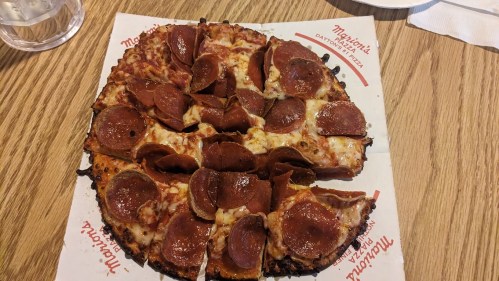 A round pizza topped with pepperoni slices, cut into triangular pieces, on a branded paper plate.
