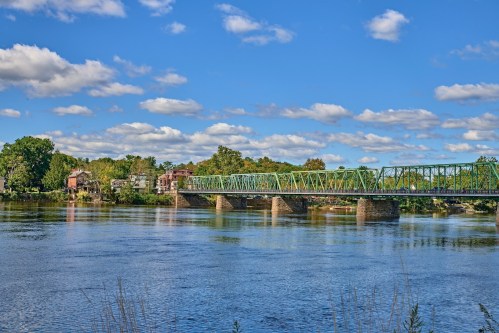 A green bridge spans a calm river under a blue sky with fluffy clouds, surrounded by trees and buildings.
