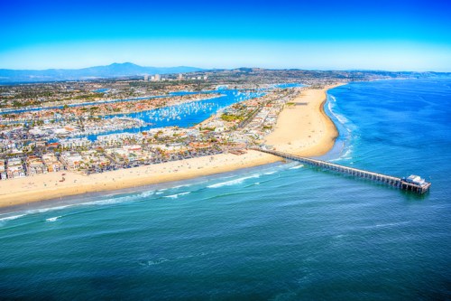 Aerial view of a coastal city with a sandy beach, marina, and pier under a clear blue sky.
