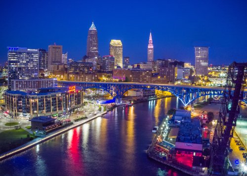 Cleveland skyline at night, featuring illuminated buildings and a bridge over the river, reflecting colorful lights.