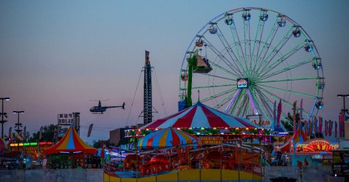 A vibrant carnival scene at dusk, featuring a Ferris wheel, colorful tents, and a helicopter in the sky.