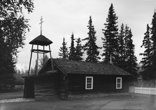 A rustic wooden church with a bell tower, surrounded by tall trees, captured in black and white.