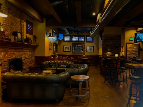 Cozy bar interior with a leather couch, brick fireplace, and wooden tables under warm lighting.