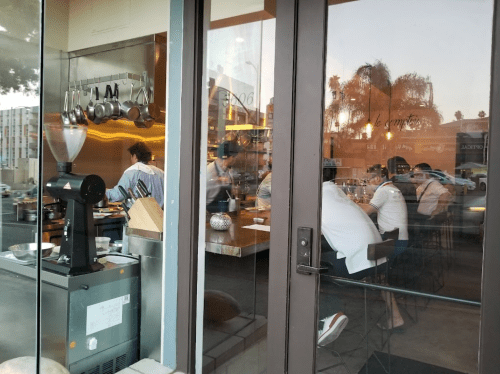 A restaurant scene viewed through glass, showing chefs at work and diners seated at a table.