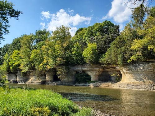 A serene river scene with rocky cliffs and lush green trees under a blue sky with scattered clouds.