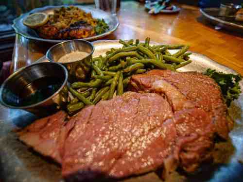 A plate of sliced roast beef with green beans and dipping sauces, served alongside a dish of salmon in the background.