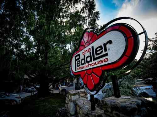 Sign for The Peddler Steakhouse, featuring a red flower design, surrounded by trees and parked cars.