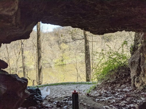 View from a cave opening, showcasing trees and a river in the background, with a backpack and water bottle in the foreground.