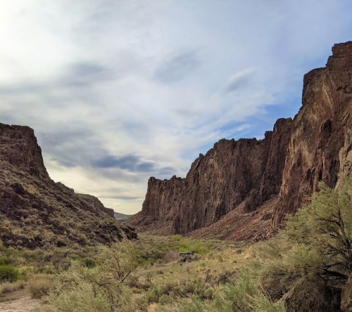 A scenic view of a canyon with steep rock formations and a cloudy sky above, surrounded by sparse vegetation.