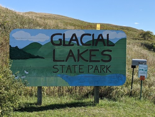 Sign for Glacial Lakes State Park, featuring green hills and a blue lake under a clear blue sky.