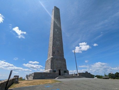 A tall stone monument under a blue sky with clouds, surrounded by grassy areas and visitors.