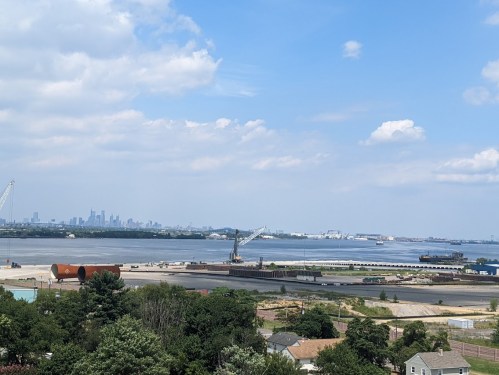A view of a waterfront with a crane, cargo ships, and a city skyline in the background under a partly cloudy sky.