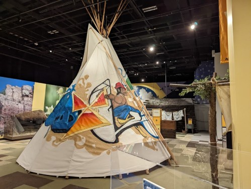 A colorful teepee with artistic designs, displayed in a museum setting with various exhibits in the background.