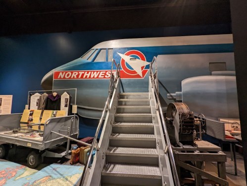 Stairs lead up to a mural of a vintage airplane with a Northwest Airlines logo in a museum setting.