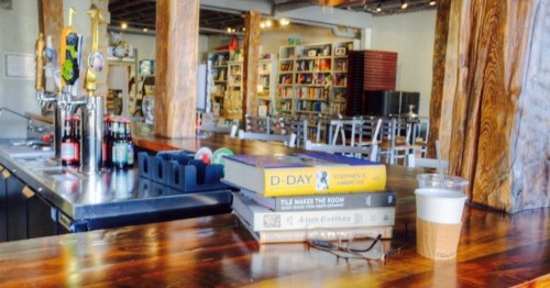 A cozy café interior with a wooden bar, stacked books, a coffee cup, and shelves filled with books in the background.