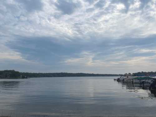 A calm lake under a cloudy sky, with boats docked along the shore and trees in the background.