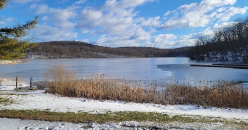 A serene winter landscape featuring a frozen lake, snow-covered ground, and distant hills under a partly cloudy sky.