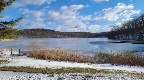 A serene winter landscape featuring a partially frozen lake, snow-covered ground, and distant hills under a blue sky.