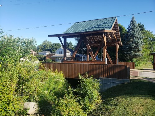 A wooden pavilion with a green roof stands near a residential area, surrounded by greenery and a clear blue sky.