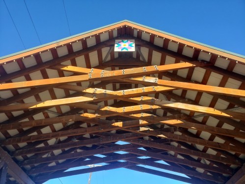 Wooden roof structure with exposed beams and a colorful star design at the peak against a clear blue sky.