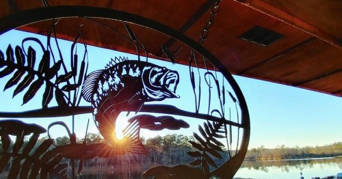 A metal fish sculpture hangs in front of a serene lake at sunset, with trees reflecting in the water.